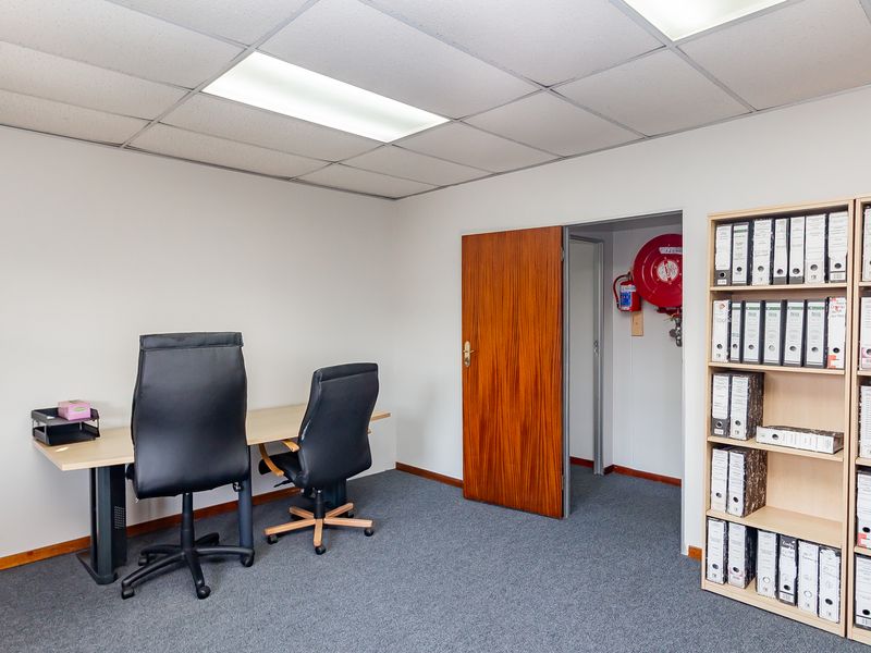 A functional office space featuring carpeted flooring, a suspended ceiling with recessed lighting, and a wall-mounted fire hose reel.