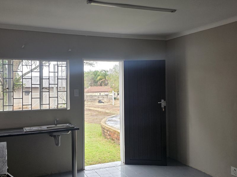 A compact, empty office kitchenette featuring a stainless steel sink, tiled flooring, and direct access to the exterior through a single door.
