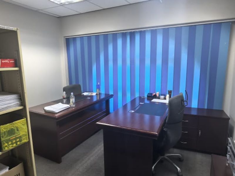 A functional office space featuring carpet flooring, a suspended ceiling with recessed lighting, and vertical window blinds.