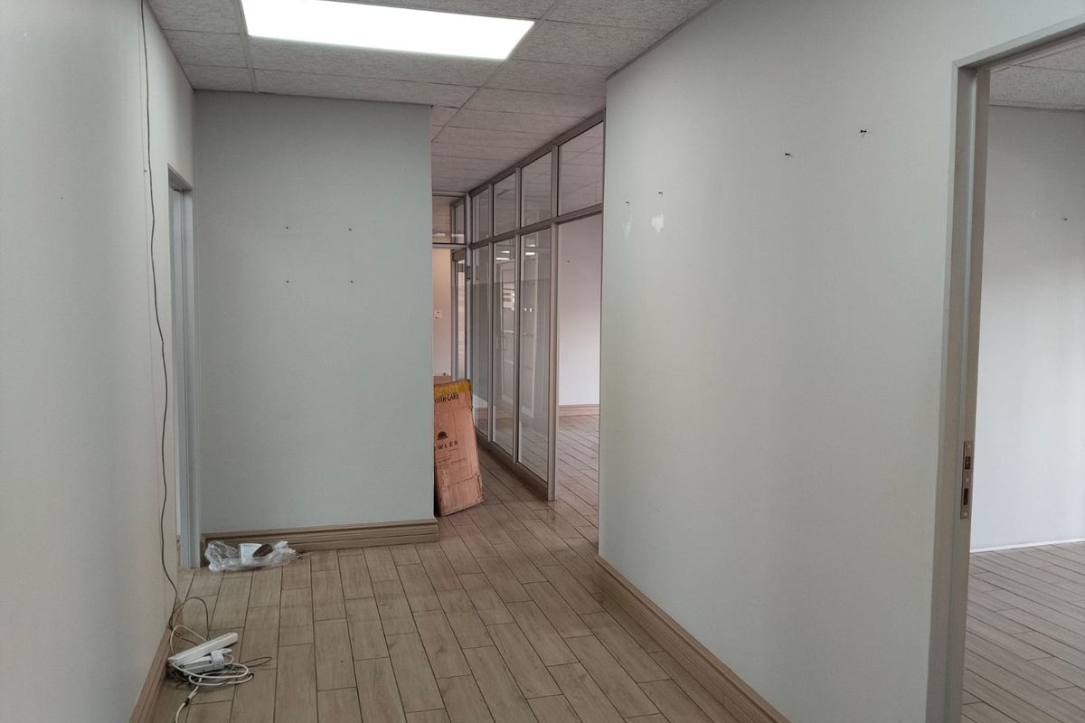 A vacant office hallway featuring wood-look tile flooring, glass partition walls, and a suspended ceiling with recessed lighting.