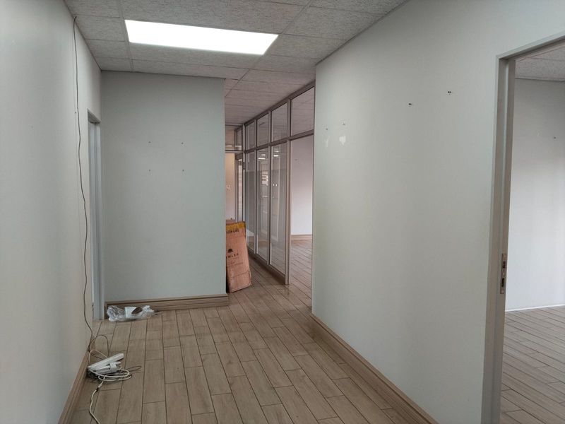 A vacant office hallway featuring wood-look tile flooring, glass partition walls, and a suspended ceiling with recessed lighting.