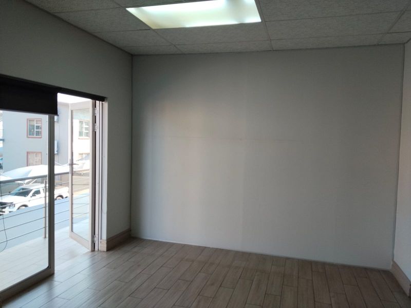 A bright, empty office space featuring modern tiled flooring, a suspended ceiling with recessed lighting, and direct access to an outdoor balcony.