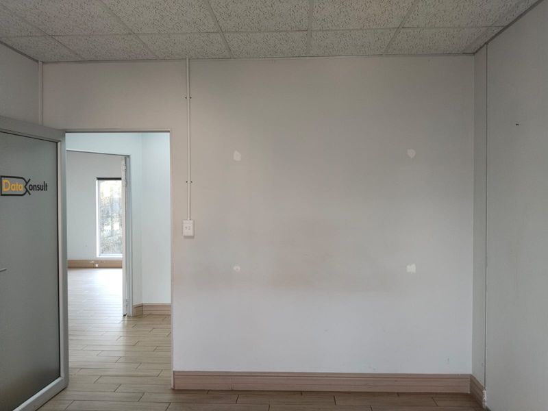 An empty office space featuring tiled flooring, a suspended ceiling, and neutral walls with visible surface patches.