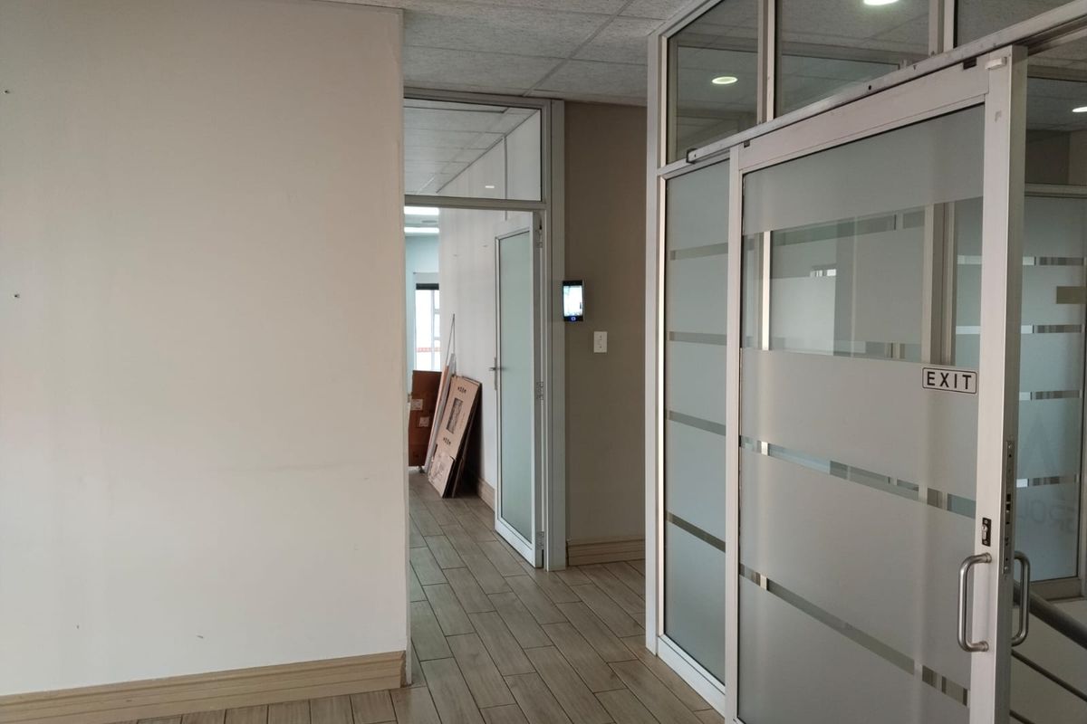 A bright office hallway featuring modern glass partitions, sliding doors, wood-look tile flooring, and a suspended ceiling with recessed lighting.