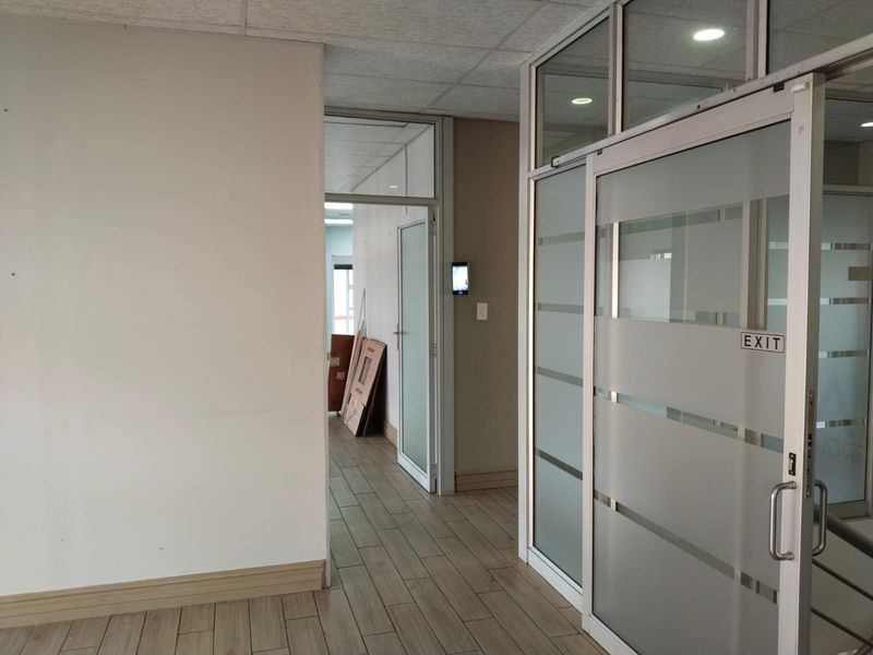 A bright office hallway featuring modern glass partitions, sliding doors, wood-look tile flooring, and a suspended ceiling with recessed lighting.
