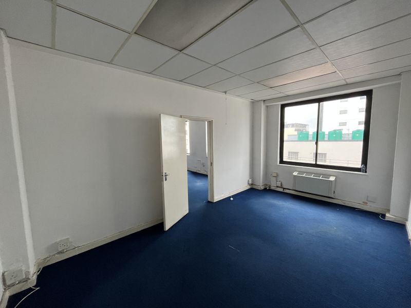 An empty office space featuring blue carpeting, a suspended ceiling, and a window-mounted HVAC unit, offering a blank canvas for professional use.