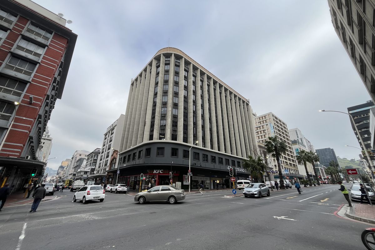 Street-level view of a multi-story commercial office building located on a busy corner, featuring ground-floor retail space and a prominent concrete facade.