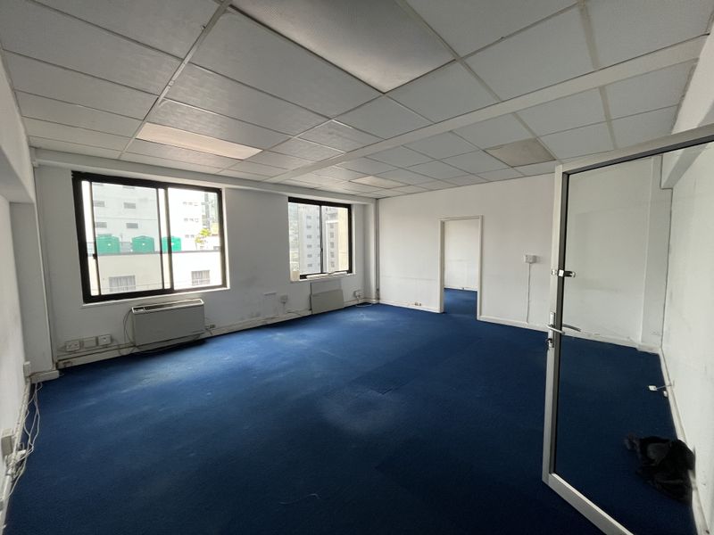 An empty office space featuring blue carpeting, a suspended ceiling, and large windows providing natural light and urban views.