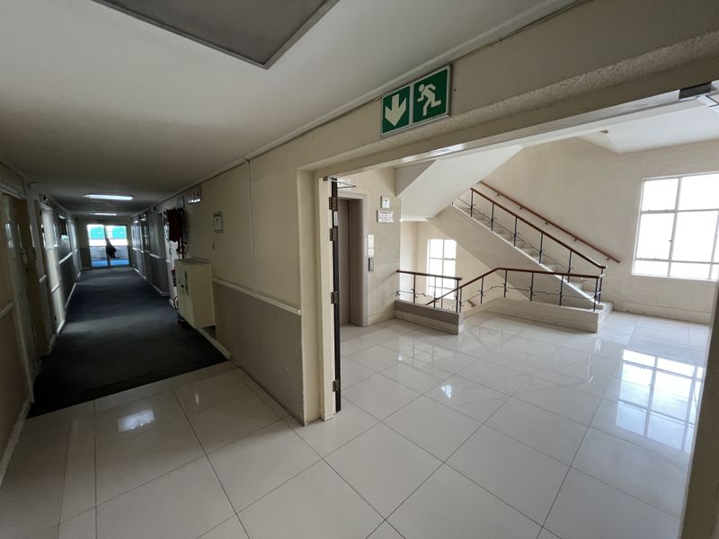 A bright office building hallway featuring tiled flooring, an elevator access point, and a staircase with a decorative wrought iron balustrade.