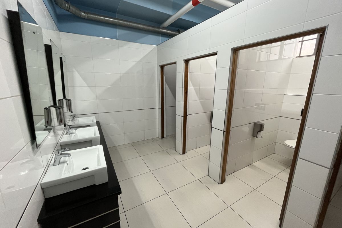 A clean, modern commercial bathroom featuring multiple vanity basins, tiled walls and floors, and private toilet cubicles with exposed industrial-style ceiling piping.