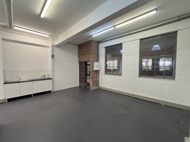 Open-plan office space featuring industrial-style steel windows, exposed brick accents, fluorescent lighting, and a compact kitchenette area with a sink.
