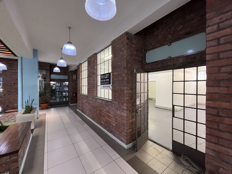 A well-maintained industrial-style hallway featuring exposed brick walls, tiled flooring, and large metal-framed glass doors leading into an open commercial space.
