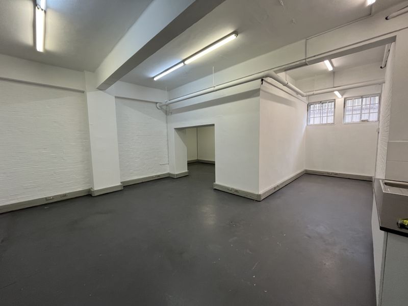 An open-plan commercial space featuring painted brick walls, industrial-style exposed ceiling pipes, fluorescent lighting, and a small built-in sink area.