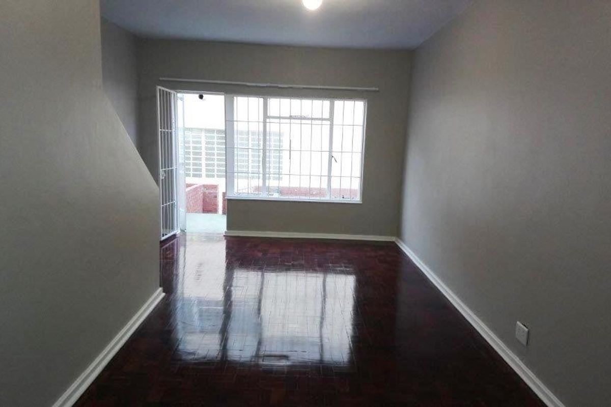 A bright, empty living room featuring polished parquet flooring, neutral walls, and a large window with security bars leading to an outdoor area.
