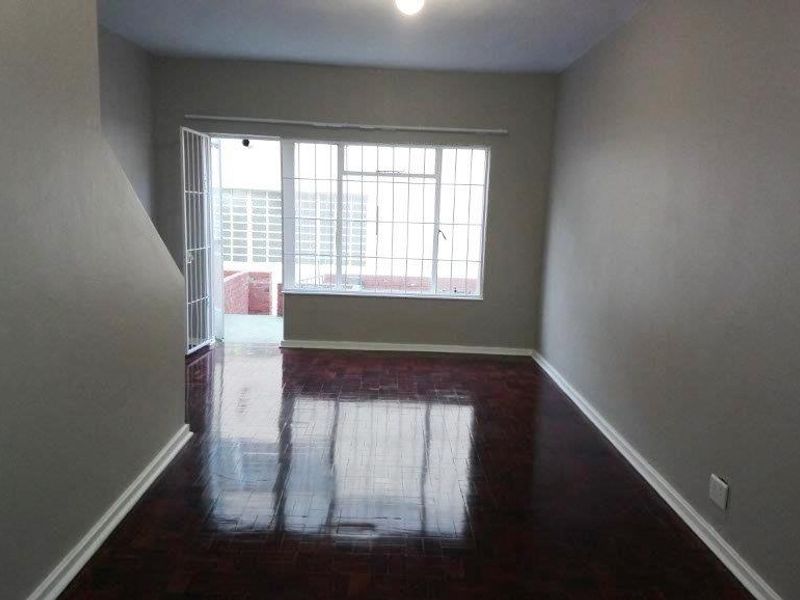 A bright, empty living room featuring polished parquet flooring, neutral walls, and a large window with security bars leading to an outdoor area.