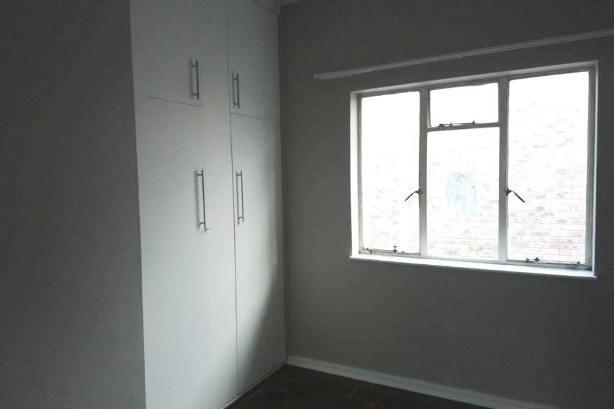 A neutral bedroom featuring built-in cupboards, parquet flooring, and a large casement window providing natural light.