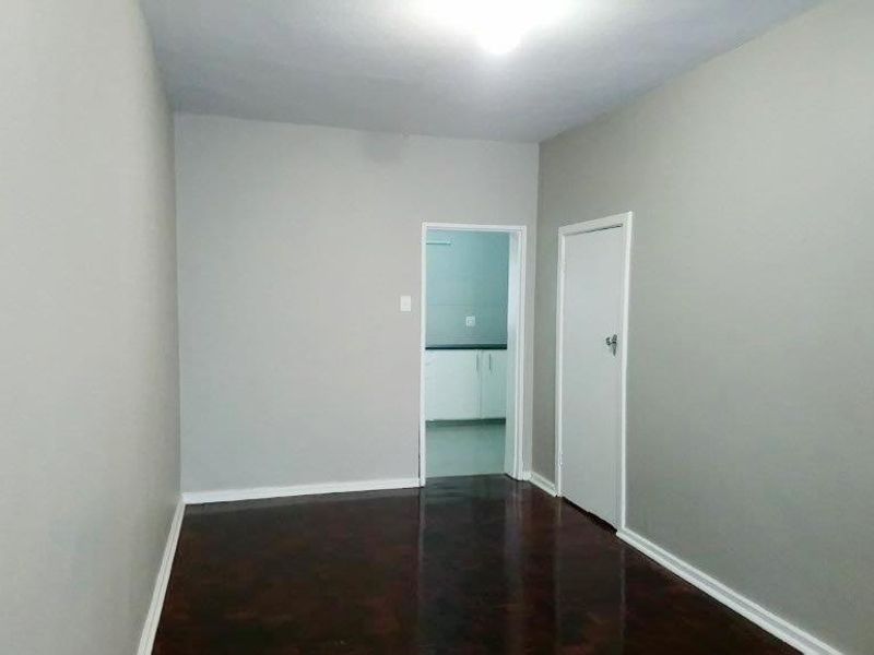An empty living room featuring dark wood-look flooring and neutral painted walls, with a doorway leading to an adjacent kitchen area.