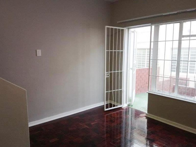 An empty living room featuring polished parquet flooring, a security gate leading to an outdoor area, and large windows providing natural light.