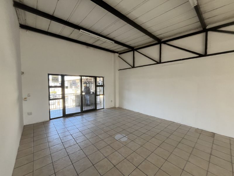 An empty open-plan commercial space featuring tiled flooring, exposed steel roof trusses, and a sliding glass door with security gate access.