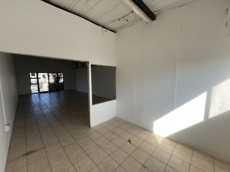 An empty, open-plan commercial space featuring tiled flooring, exposed ceiling beams, and an internal partition wall with a pass-through window.