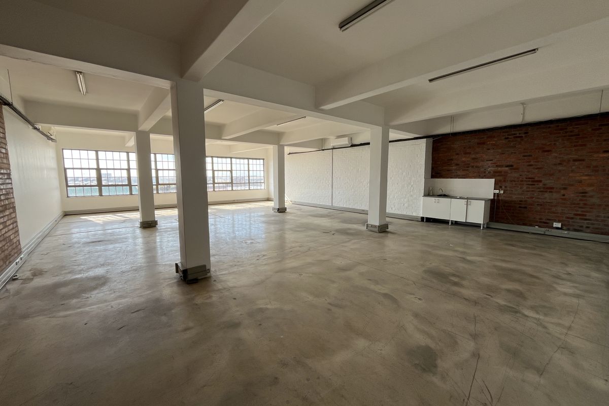 Spacious open-plan industrial office space featuring exposed brick walls, polished concrete floors, large windows, and a compact kitchenette unit.