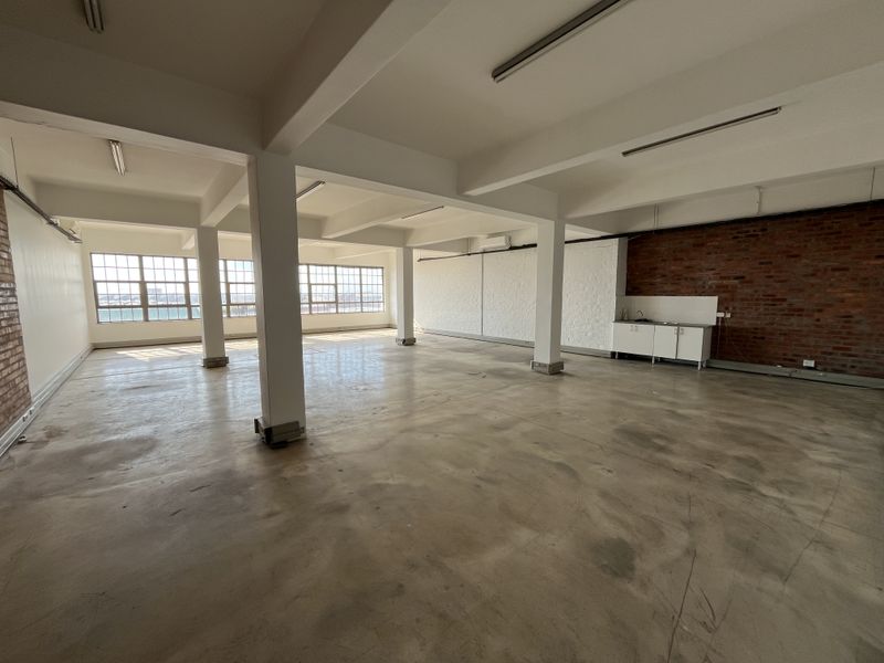 Spacious open-plan industrial office space featuring exposed brick walls, polished concrete floors, large windows, and a compact kitchenette unit.