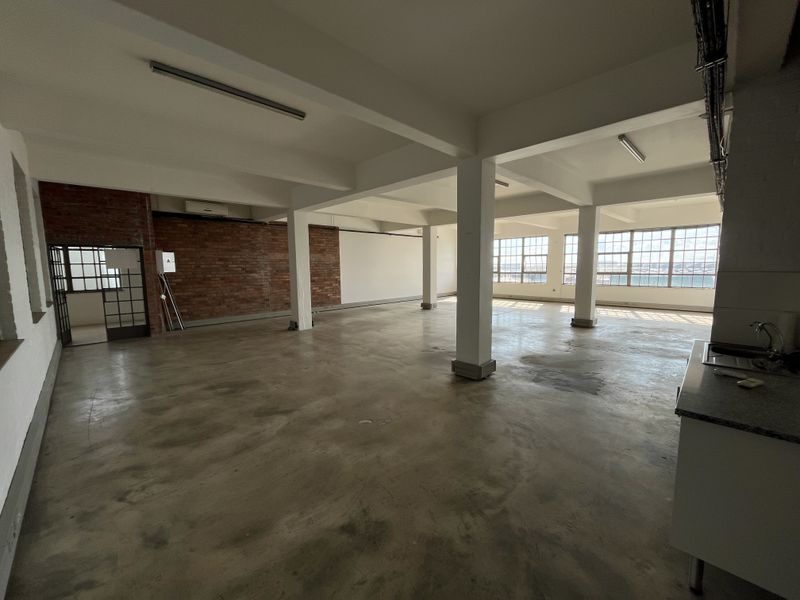 Spacious open-plan industrial office space featuring exposed brickwork, polished concrete floors, large steel-framed windows, and a functional kitchenette area.