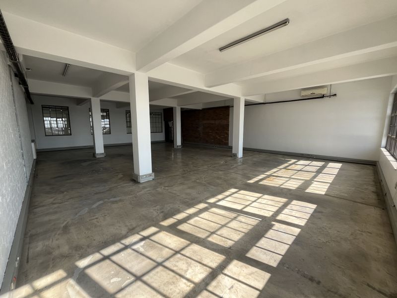 Spacious open-plan industrial office space featuring polished concrete floors, exposed structural columns, brick accents, and large steel-framed windows providing ample natural light.