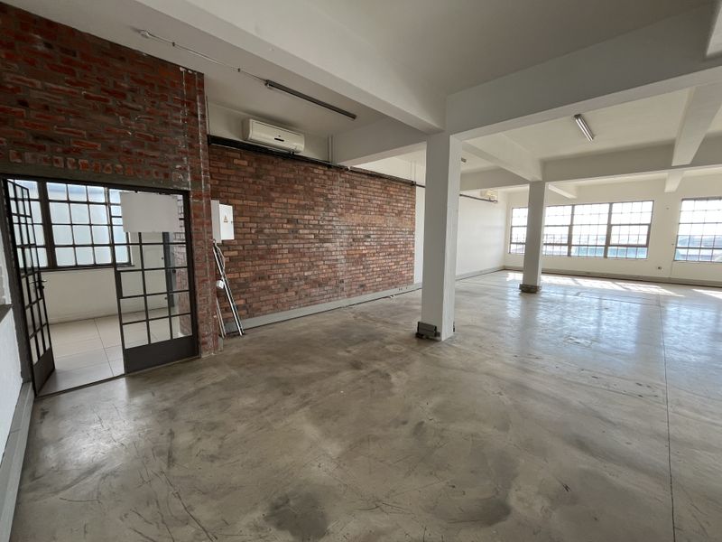 Spacious open-plan commercial office area featuring industrial-style exposed brick walls, polished concrete flooring, and large steel-framed windows providing ample natural light.