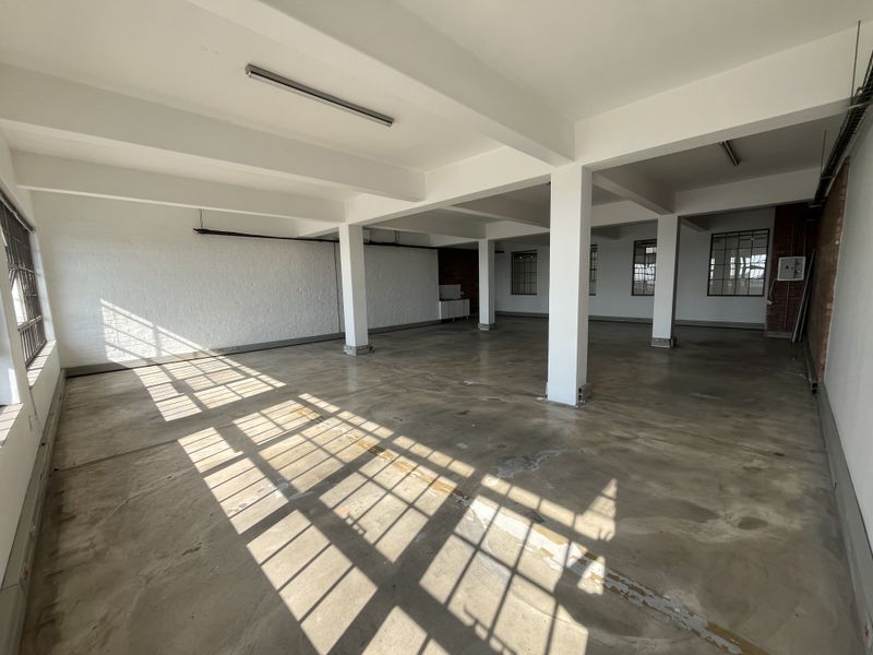 Spacious open-plan commercial office area featuring industrial-style concrete flooring, exposed structural columns, and large steel-framed windows providing ample natural light.
