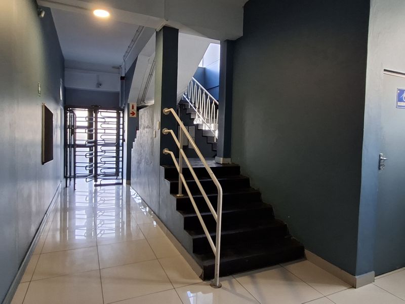A clean, well-lit apartment building hallway featuring tiled floors, a staircase with a metal handrail, and a security turnstile at the entrance.