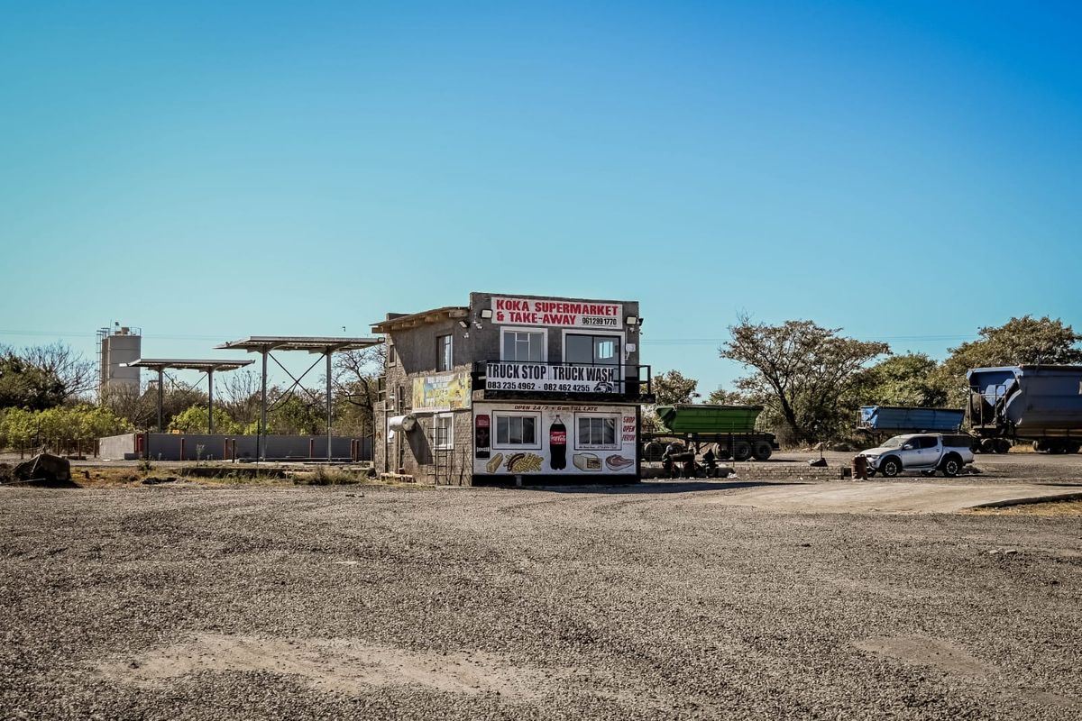 Exterior view of a commercial truck stop facility featuring a two-story building with a supermarket, take-away, and truck wash services on a gravel lot.