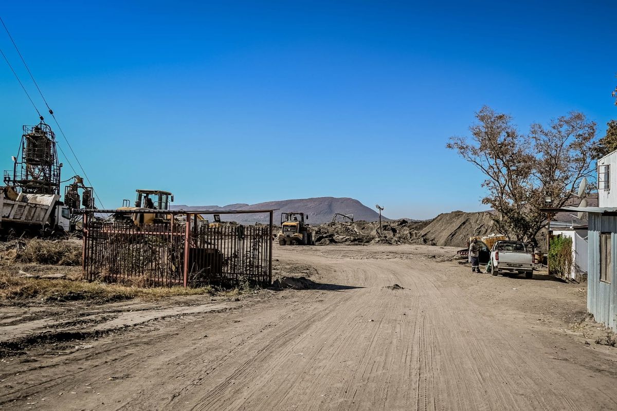 An industrial outdoor site featuring an unpaved dirt road, metal fencing, and heavy machinery in a rural setting.