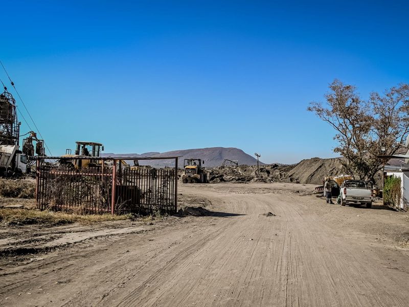 An industrial outdoor site featuring an unpaved dirt road, metal fencing, and heavy machinery in a rural setting.