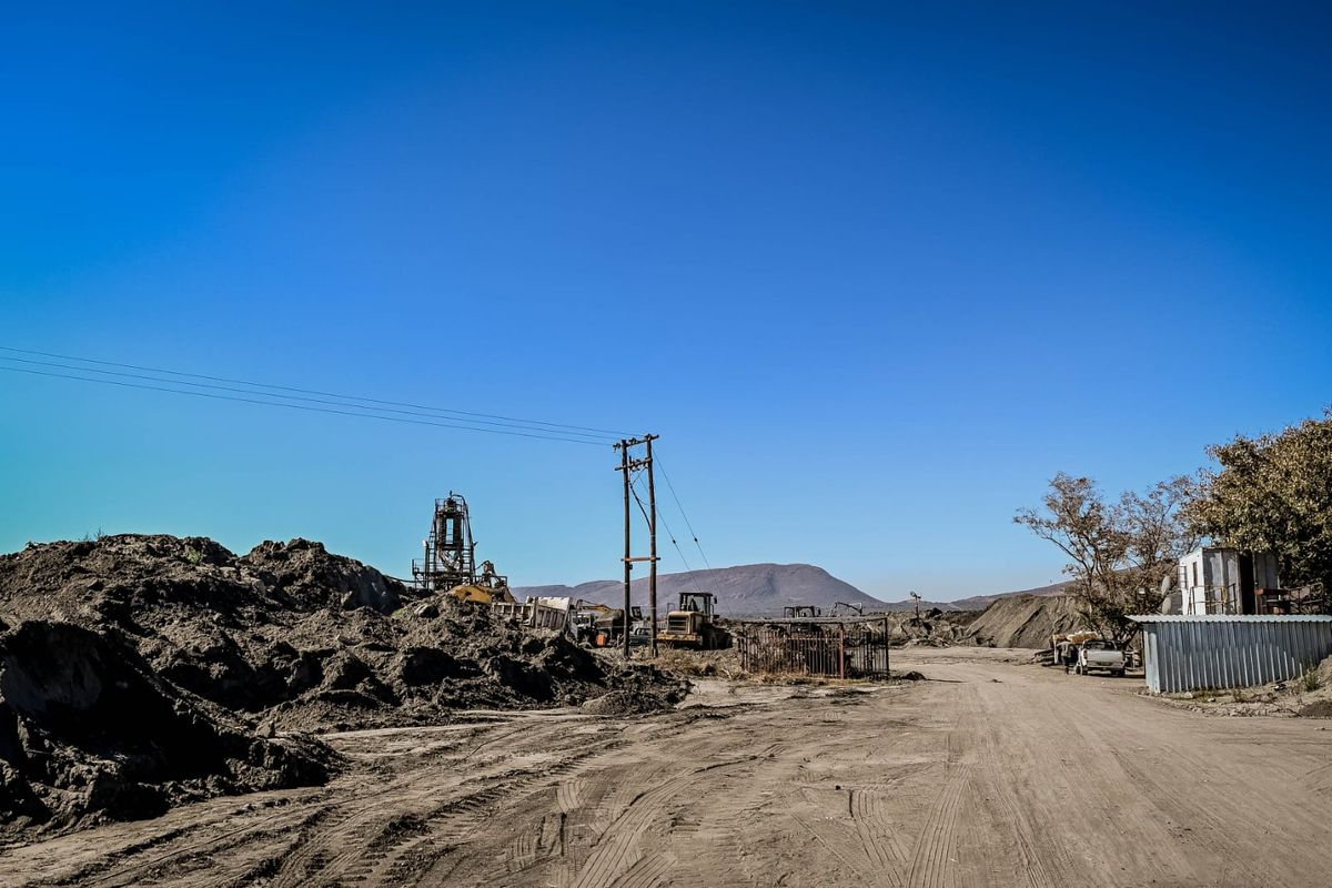 An industrial site featuring unpaved ground, utility infrastructure, and a corrugated metal structure set against a mountainous landscape.