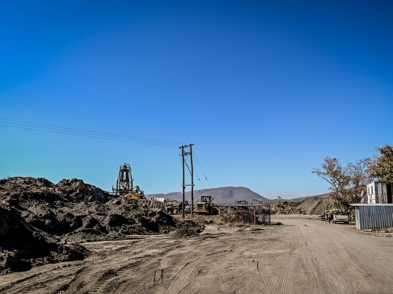 An industrial site featuring unpaved ground, utility infrastructure, and a corrugated metal structure set against a mountainous landscape.