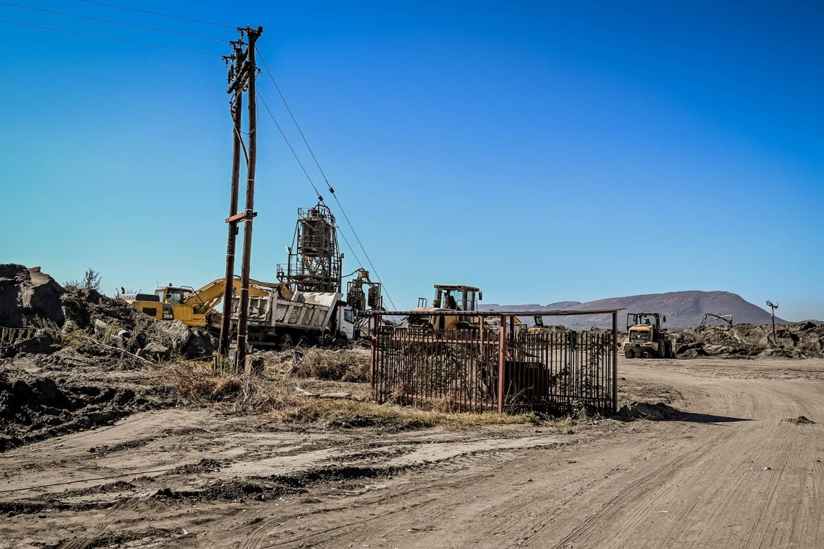 An industrial site featuring heavy machinery, earthworks, utility poles, and metal fencing in a rural, open-air setting.