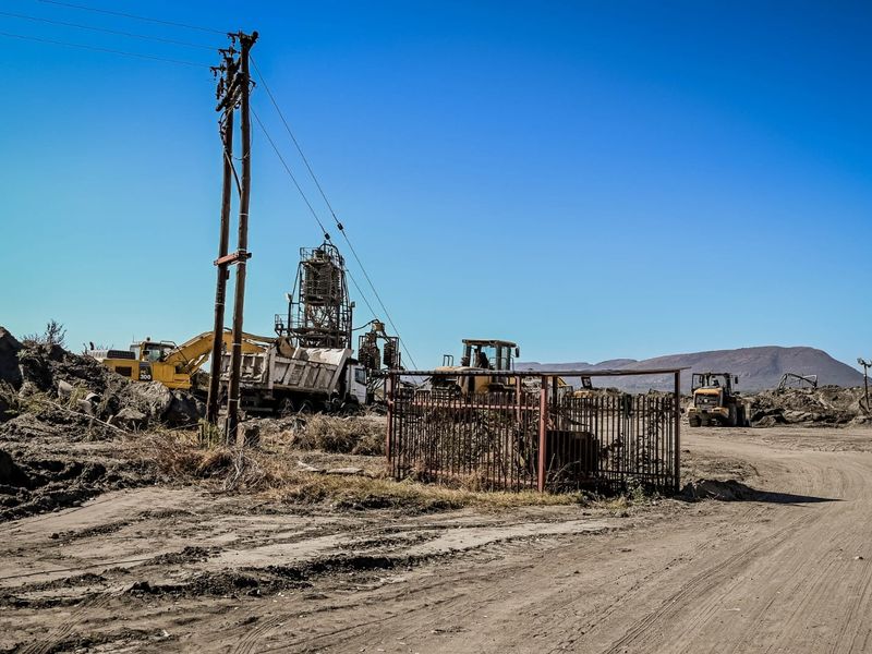 An industrial site featuring heavy machinery, earthworks, utility poles, and metal fencing in a rural, open-air setting.