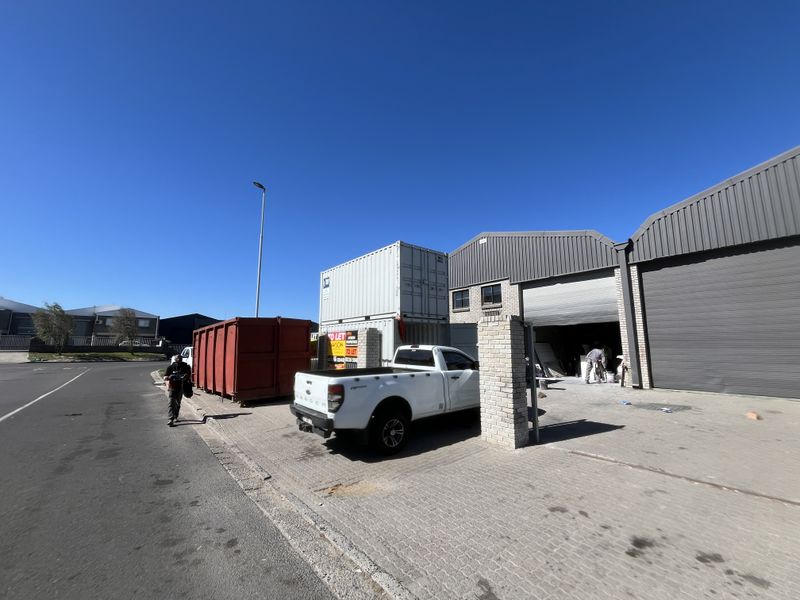Exterior view of an industrial warehouse property featuring metal cladding, roller shutter doors, and a paved driveway area.