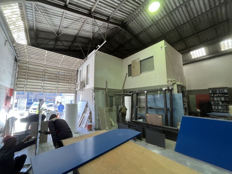 Industrial warehouse space featuring high ceilings with exposed steel trusses, a mezzanine level, concrete flooring, and a large roller shutter door for vehicle access.