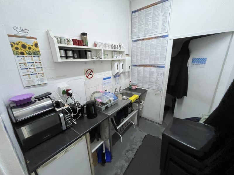 A compact staff kitchenette featuring a stainless steel sink, wall-mounted shelving, and tiled splashback, requiring cosmetic updates to improve functionality and appearance.