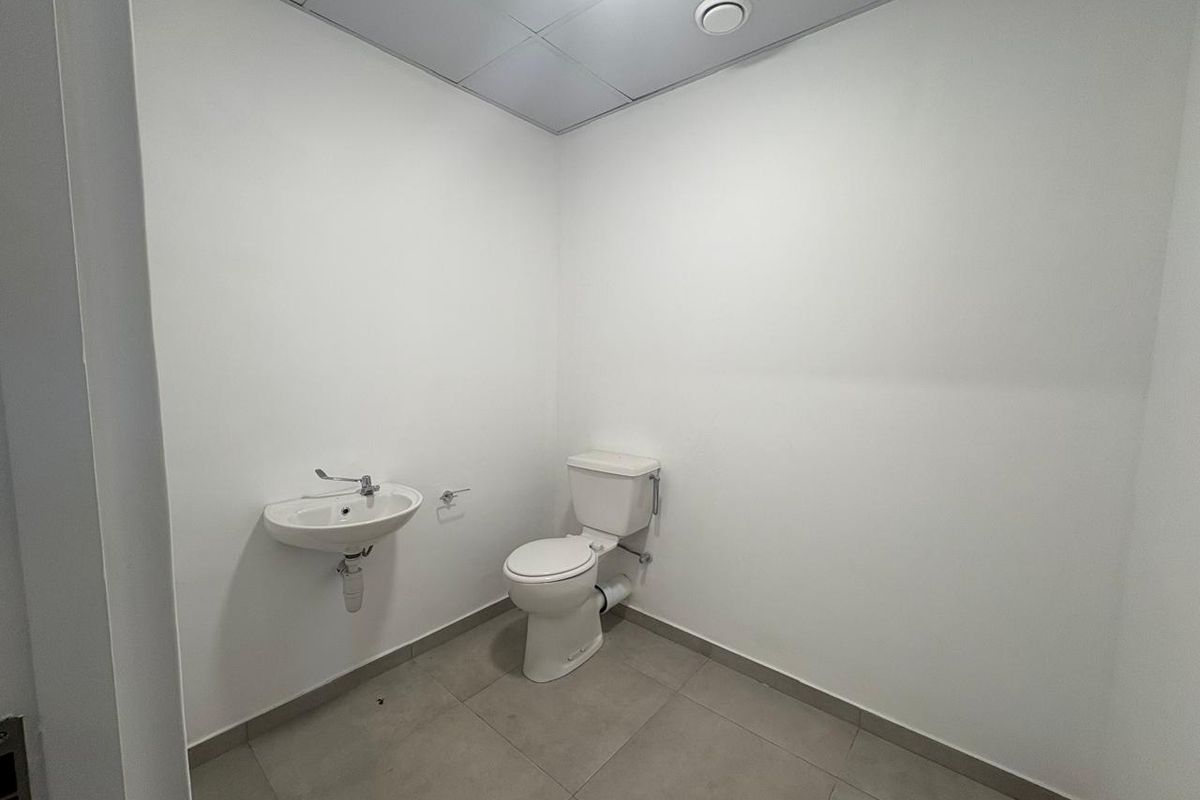 A clean, minimalist industrial-style bathroom featuring a wall-mounted basin, standard toilet, tiled flooring, and a suspended ceiling.
