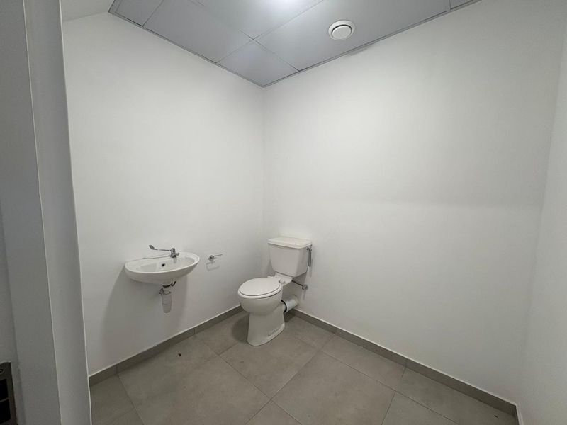 A clean, minimalist industrial-style bathroom featuring a wall-mounted basin, standard toilet, tiled flooring, and a suspended ceiling.