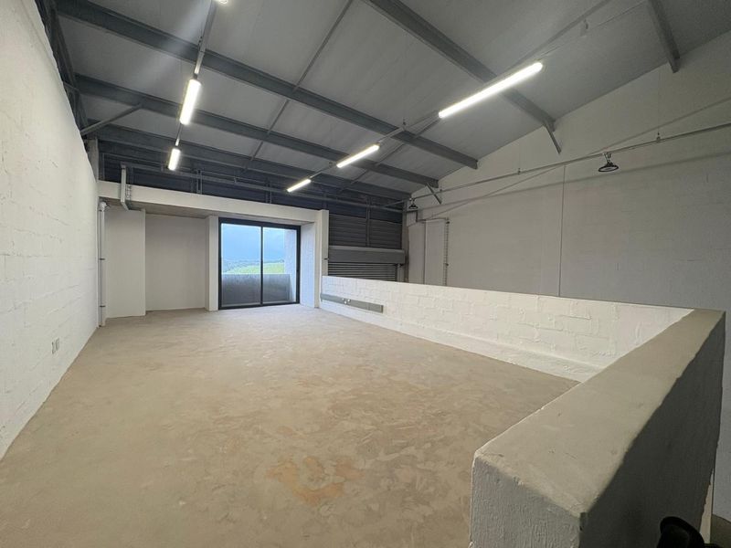 Spacious open-plan industrial mezzanine area featuring exposed steel roof trusses, concrete flooring, and natural light through a sliding glass door.