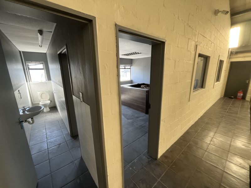 A functional industrial hallway featuring tiled flooring, exposed brick walls, and access to adjacent office and restroom facilities.