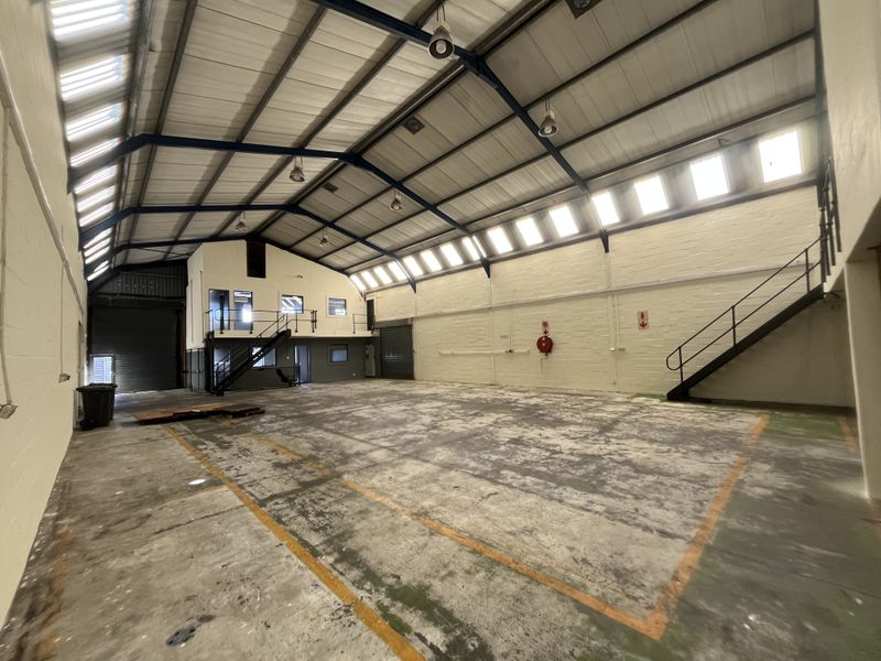 Spacious industrial warehouse featuring high ceilings with exposed steel trusses, a mezzanine level with staircase, roller shutter doors, and durable concrete flooring.