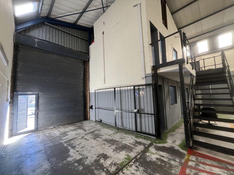 Industrial warehouse space featuring a large roller shutter door, mezzanine level with metal staircase, and high ceilings with exposed steel trusses and skylights.