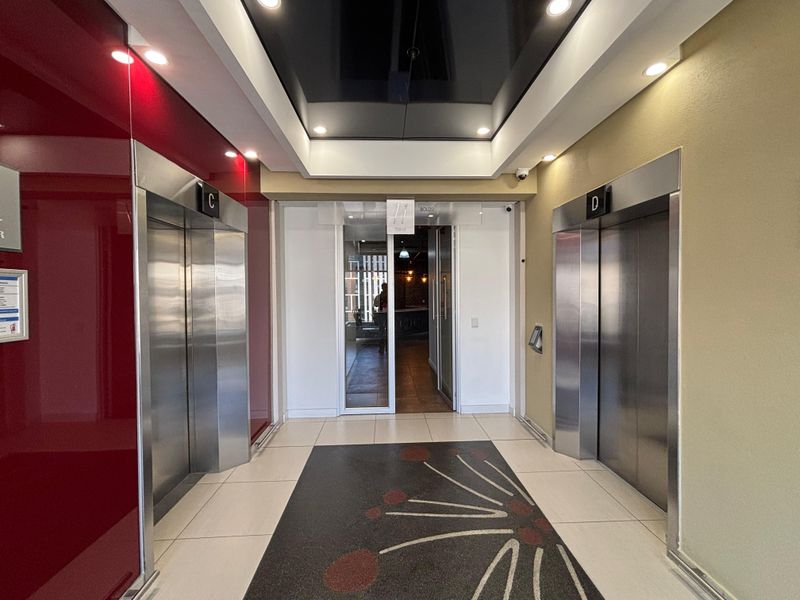 A modern office building elevator lobby featuring stainless steel lift doors, recessed ceiling lighting, and a clean, professional tiled floor finish.