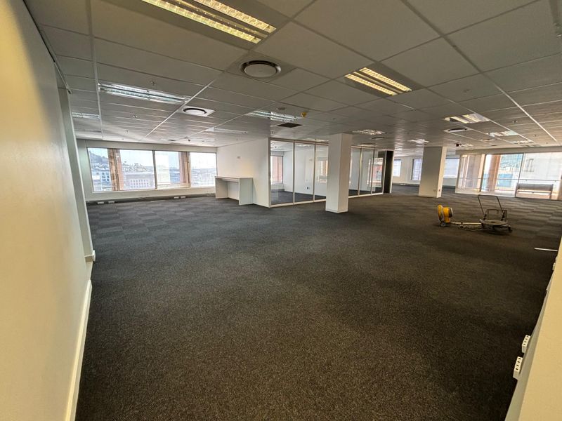 Spacious open-plan office area featuring suspended ceilings, recessed lighting, carpeted flooring, and large windows providing ample natural light and city views.