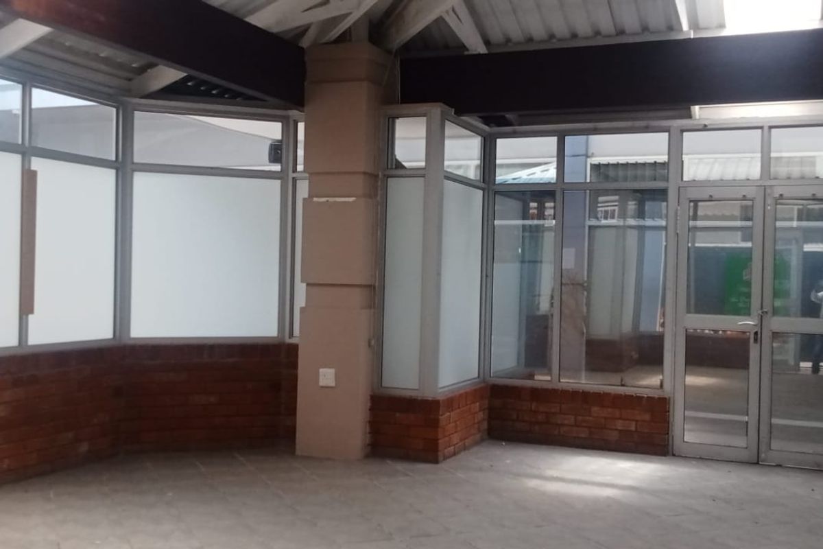 A bright, empty retail entrance area featuring exposed brickwork, large aluminium-framed glass windows, and tiled flooring, suitable for a professional storefront or reception space.